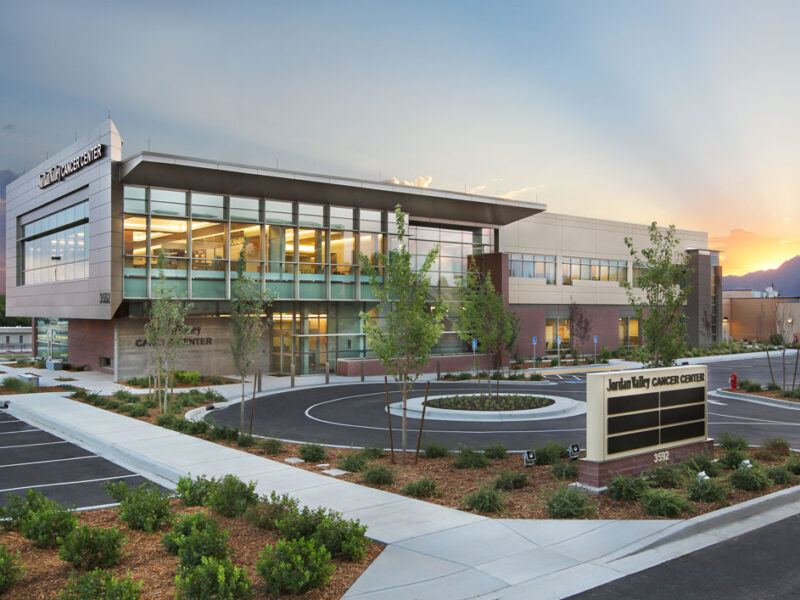 External photo of Jordan Valley Cancer Center at sunrise. View of building with parking lot in front and sun peaking over mountains in the distance. Designed by TSA Architects.
