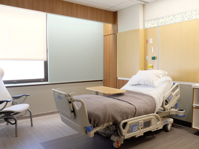 Hospital design of patient room at University of Utah. Patient bed in the middle of the room with guest chair at the end of the bed near window on far wall.
